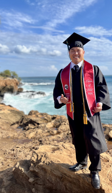 A guy is standing on a rock, throwing his "Shaka," wearing his graduation cap and gown, BSN stole, and cords with a beautiful ocean view behind him.