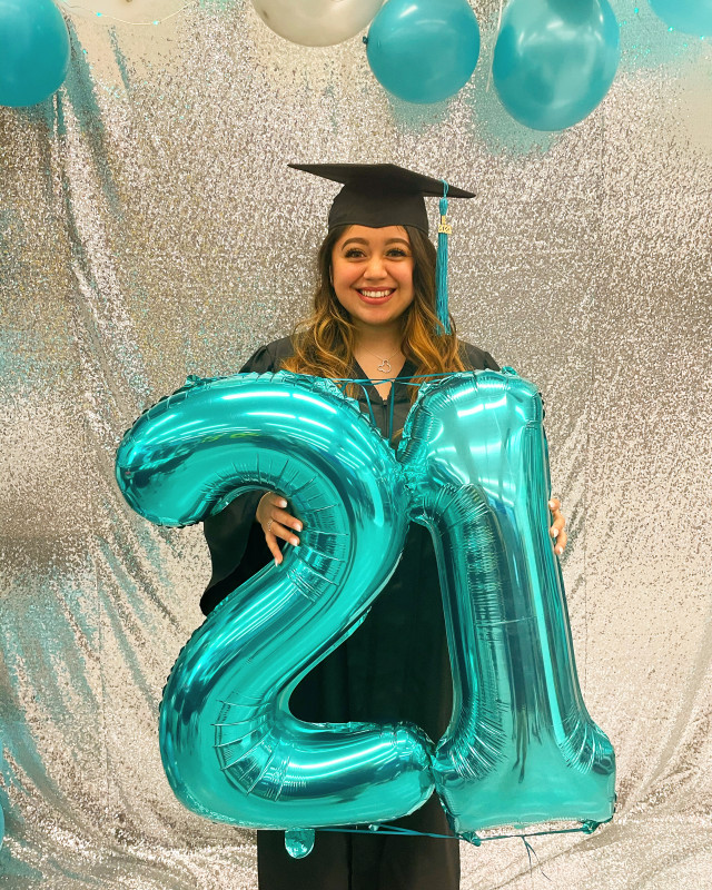 I am smiling and I have brown curly hair. I am wearing my black cap and gown.I am holding the number 21 balloons. There is a silver background with white and teal balloons.