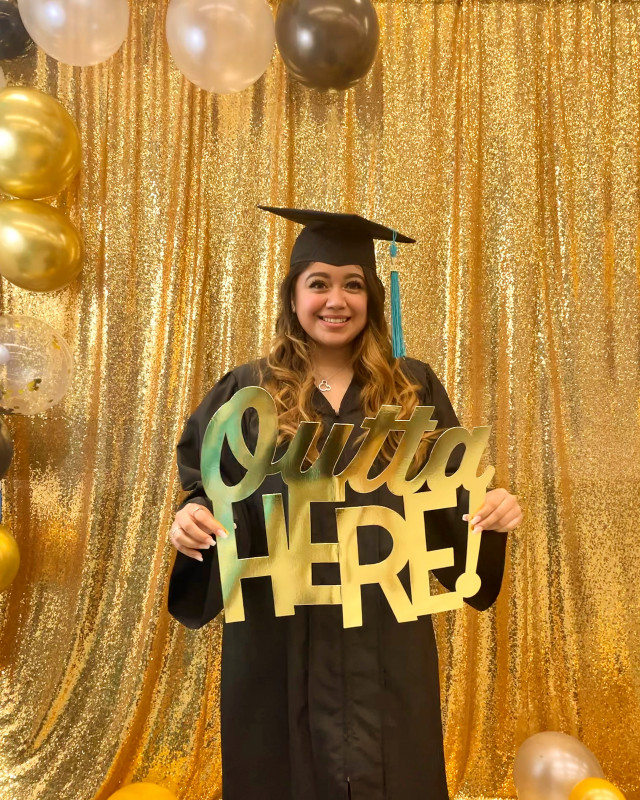 I am wearing my black cap and gown. I am holding the “outta here” gold sign. There are either and gold balloons in the background.