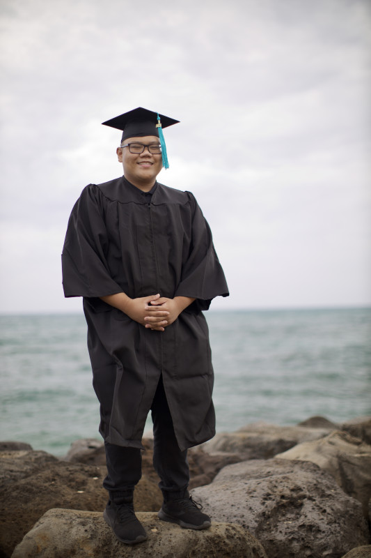 Dennis in his graduation cap and gown, on top of rocks, with a view of the ocean in the back.