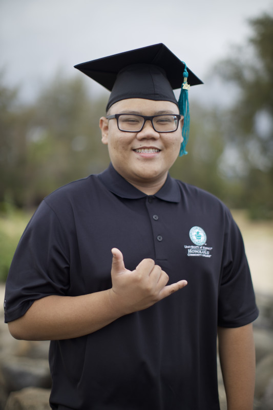 Dennis with his graduation cap, and HCC polo shirt, throwing up a Shaka.