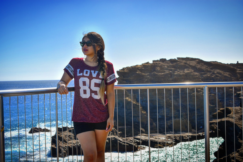 I am wearing a t-shirt and black shorts. I have my sunglasses on and looking over to my right. I am leaning against the rails and you can see the ocean behind me.