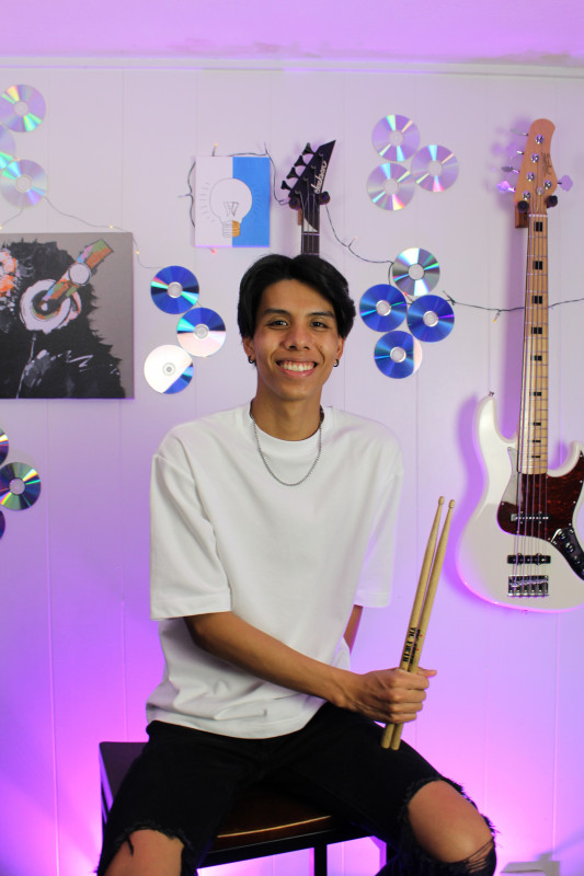 A smiling portrait of me in a studio sitting on a chair with my drumsticks in my hand.