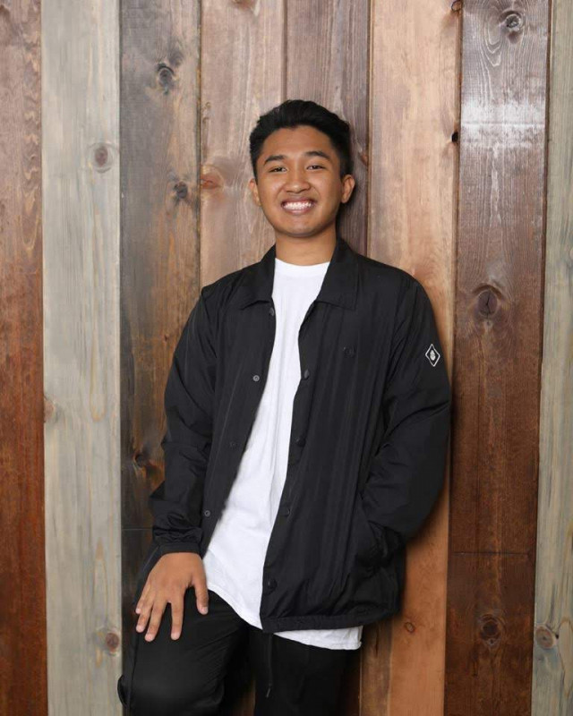 This is a 75% body shot of myself.  My right foot is resting against a wooden wall, and my right hand is resting against my pant leg.  I am smiling directly at the camera; wearing my white shirt and black windbreaker over the shirt.