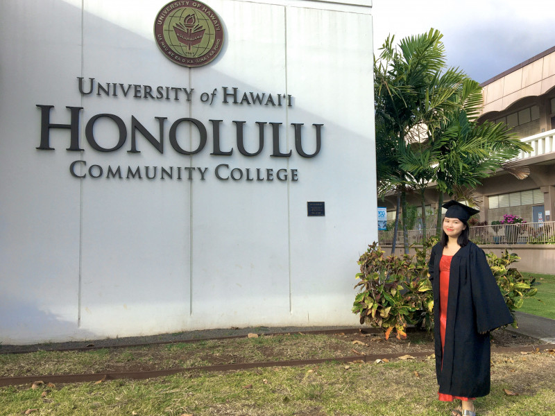A person wearing a cap, gown, and a bright red dress, standing in front of the Honolulu Community College sign by the Admin Office, but like, landscape view.