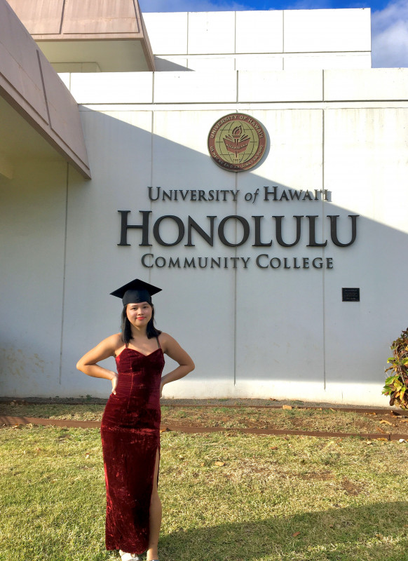 A person in a dark red dress and a cap, standing in front of the Honolulu Community College sign by the Admin Office.