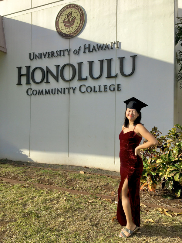 A person in a dark red dress, standing in front of the Honolulu Community College sign by the Admin Office, smiling but with one eye closed because the sun was too bright