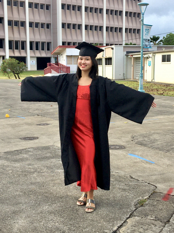 A close up shot of a smiling person wearing a cap, gown, and a bright red dress. She's standing in the main mall with Building 2 in the background, extending her arms like she's about to hug you but she's actually just showing off how big this gown is.