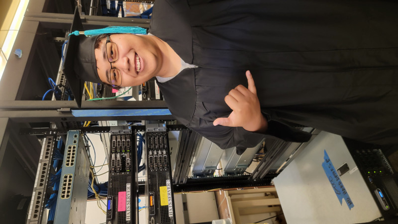 Isaiah Kaleikini wearing cap and gown giving a shaka in front of server computer.