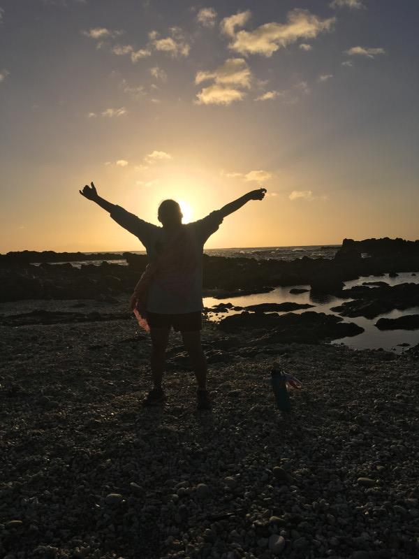 A woman jumps holding up her arms in joy at the shoreline. Behind her is a the setting sun.