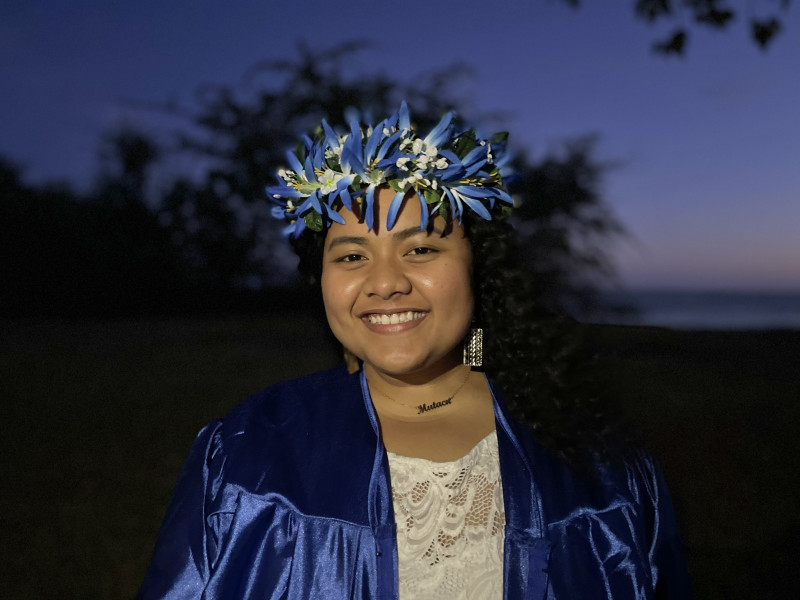 A brown woman with black curly hair facing the camera smiling wearing a white dress under a blue graduation gown. Also wearing a haku on her head with blue and white flowers.