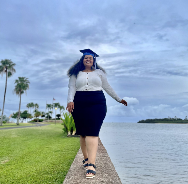 A brown woman with black curly hair standing on a rock wall and is facing the camera smiling and is wearing a white long-sleeved blouse with brown buttons on it and a black skirt. She is also wearing a blue graduation cap and tassel on her head as well.