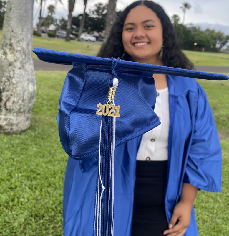A brown woman with black curly hair facing the camera smiling and holding a a graduation cap with a tassel with the year 2021 on it in focus and towards the camera, while the woman in the picture who is wearing a white dress under a blue graduation gown, is not in focus.