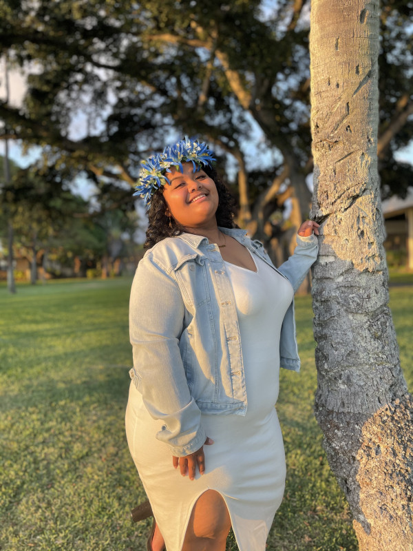 I am wearing a white dress with a Jean jacket with a blue flowered haku while leaning on a coconut tree outside.