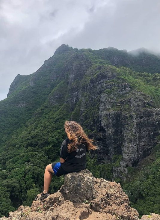 Anna sits on a rock, back to the camera because she didn't know this was being taken.  She faces a towering, dark green mountain with a grayish cloudy sky above her. The wind is blowing her hair.