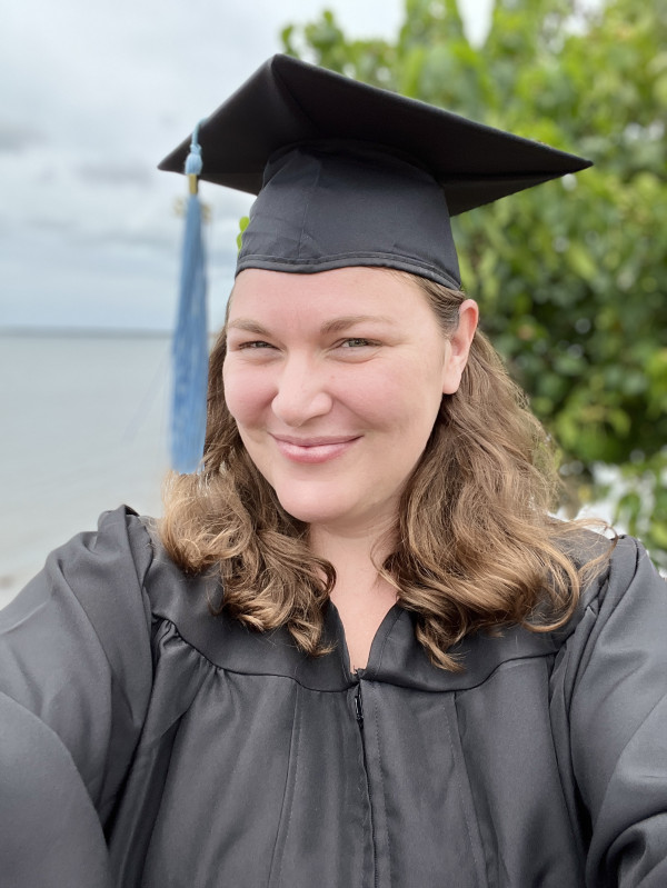 Close up of female in graduate cap and gown, shoulder length brown hair, water and tree in the background