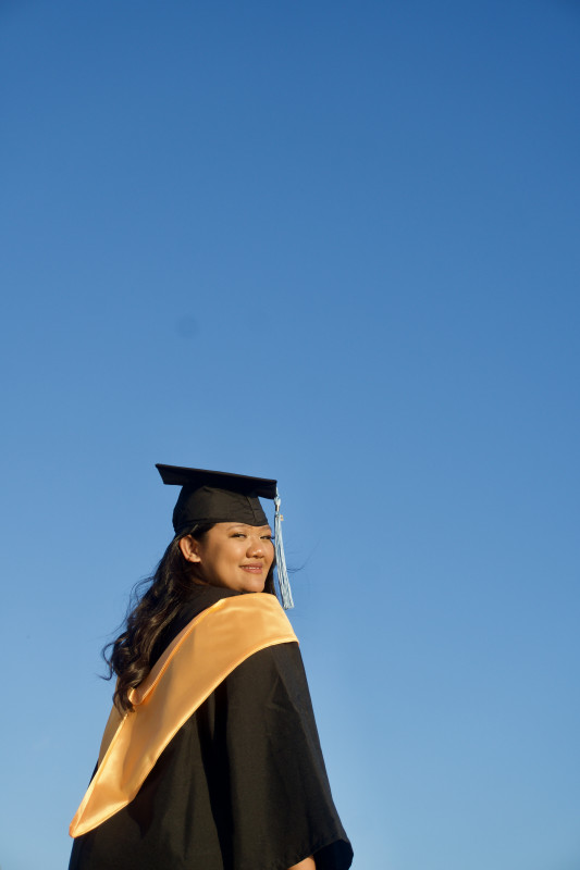 #1: A picture with my cap, gown, & 2022 tassel. This picture is take by the beach where Im standing facing on my right side. The picture also shows the blue sky as my background.