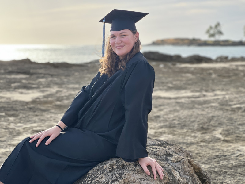 Female graduate in cap and gown sitting on a large rock with ocean in the background with tree in the distance out of focus.