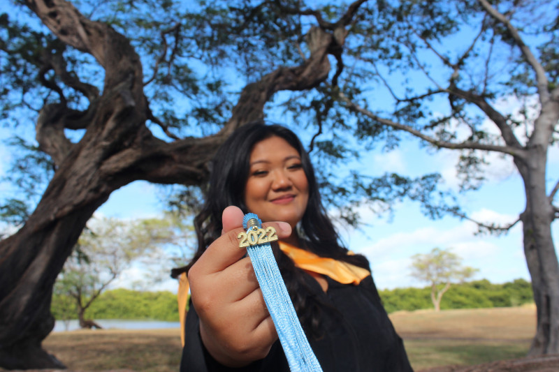 This picture is a frontal view and its me holding my 2022 tassel with my right hand. The photo emphasizes the tassel and the tree branches as the background.