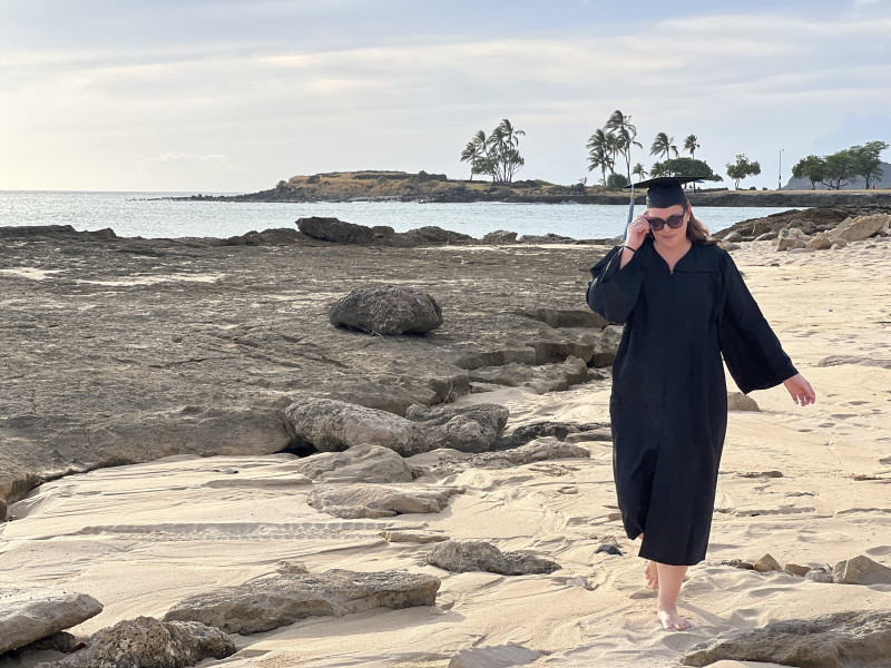 Female in graduate cap and gown and sunglasses, walking barefoot at the beach