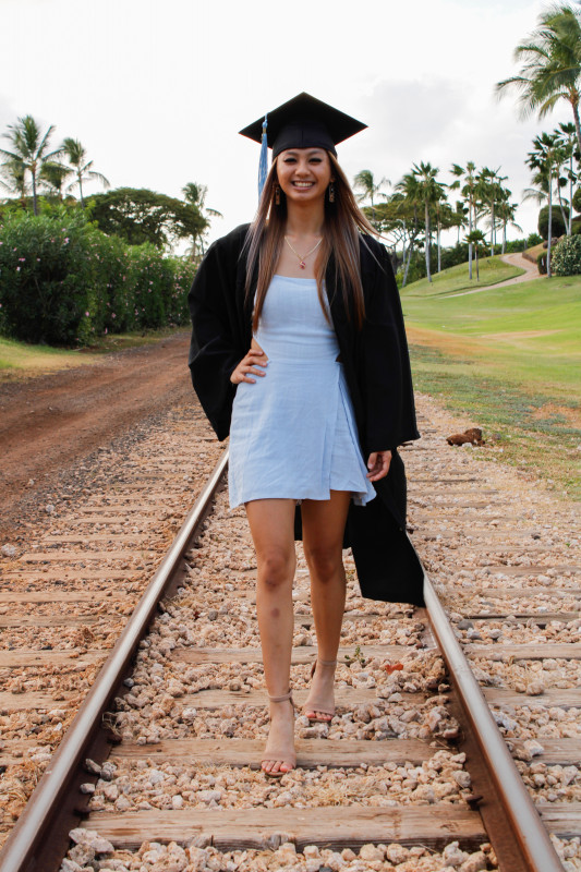 graduate with her cap and gown posing in the middle of the train tracks at Ko'olina