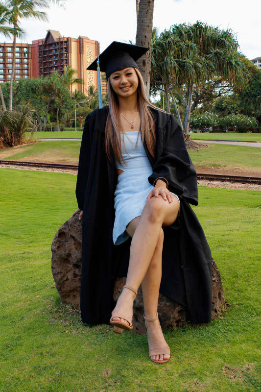 graduate with her cap and gown sitting on a rock near the train tracks at Ko'olina