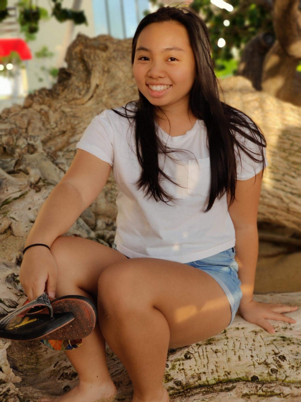 A teenage girl sitting in the shade on the beach with her slippers off and the wind lightly blowing her long hair.