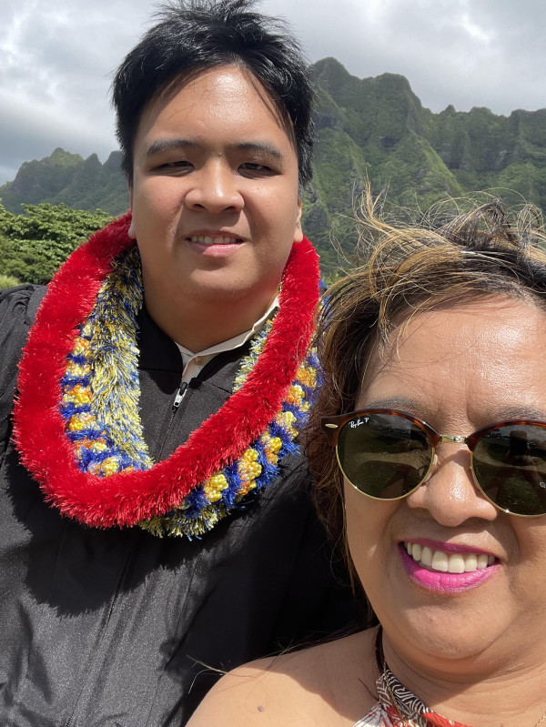 Genesis and his Mother posing in front of a green mountain in Kualoa Park in Graduation garb.