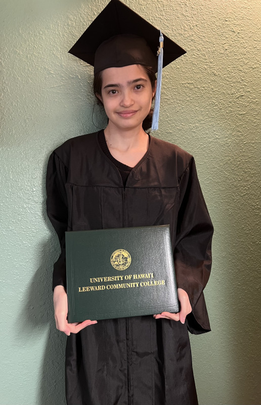 A Leeward Community College Graduate Student who is wearing a black dress with a black and cap and gown holding a Leeward Community College degree.