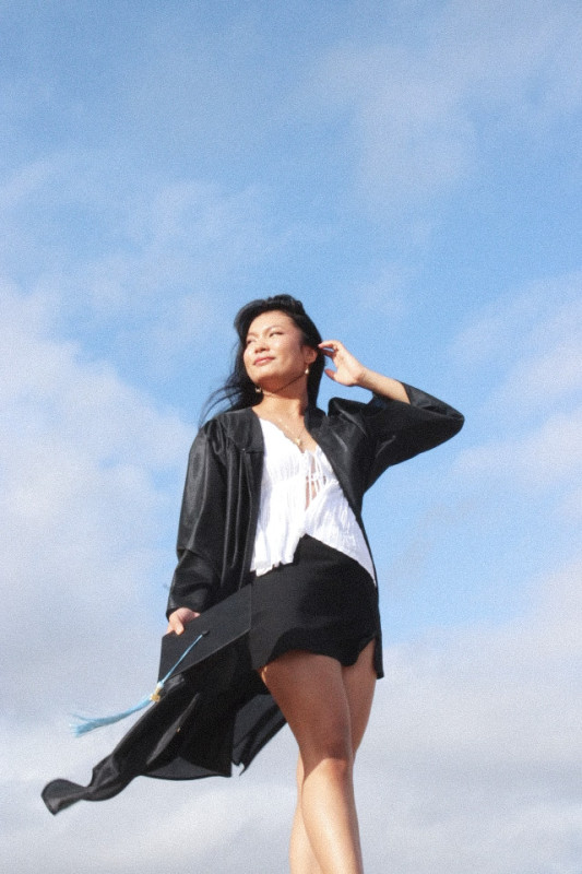 The graduate in the center with the blue sky behind and gray clouds.  She is wearing a black graduation gown and holding out a black cap out with her left hand. Her right hand is touching her hair, while she looks off to the left.