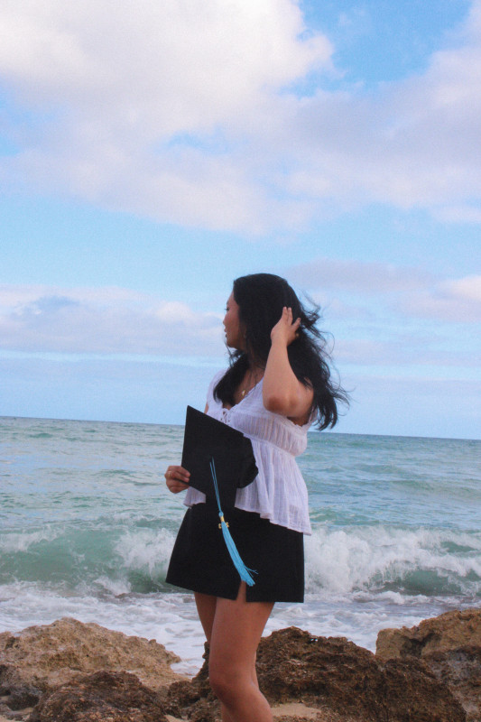 The graduate in the center looking behind her while her right arm is up by her hair. She is wearing a white top and black skirt. The background is a blue sky with the ocean, and there is brown rocks at the bottom.