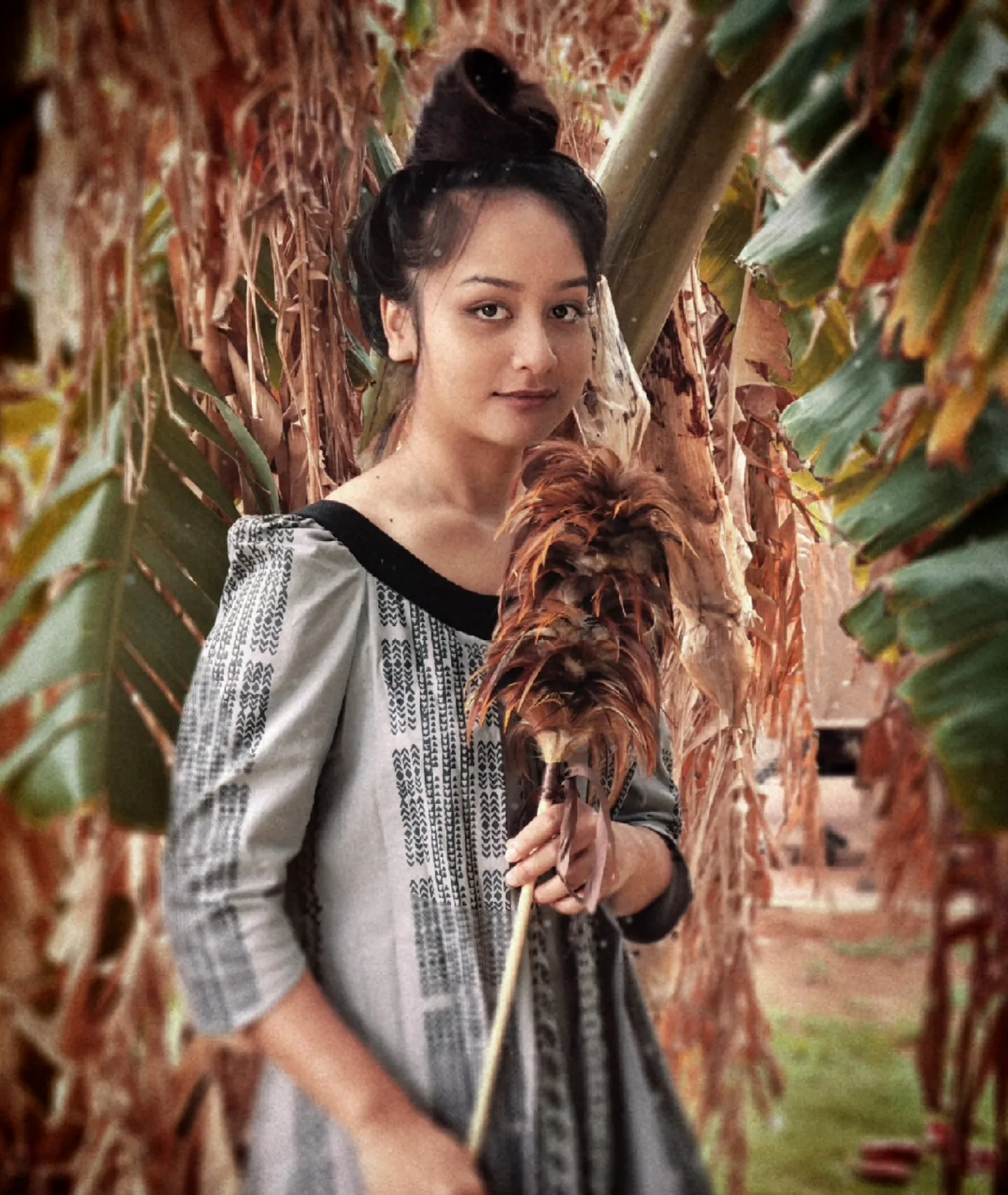 This image is a picture of a lady dressed in a muʻumuʻu(traditional kupuna wear), that is colored grey and black.  She holds a kahili on with hear left hand holding the tip of the stick and her right hand holding the lower end of the stick.  Her hair and makeup is brown and fragrant like the lush full banana trees in the back.