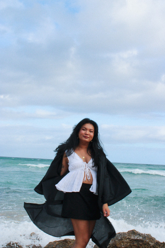 The graduate in the center, with the ocean and sky behind. The graduate is wearing a white top and black skirt, with a black graduation gown blowing out.