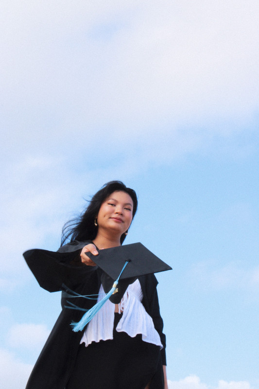 The graduate in the center, with a blue sky and clouds in the background. The graduate is wearing a white top and black skirt with a black graduation gown on, and holding out a black graduation cap with blue tassel.