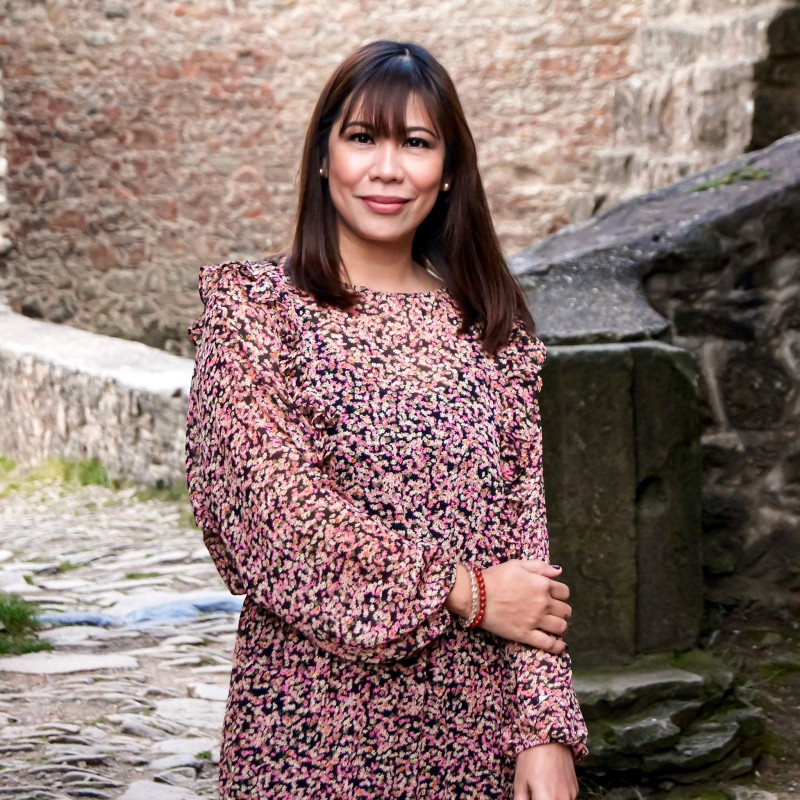 A photo of myself in a floral dress with a mixture of black, pink, and orange colors of tiny flowers. I have dark brown shoulder-length hair, bangs, brown eyes, and medium-brown skin.