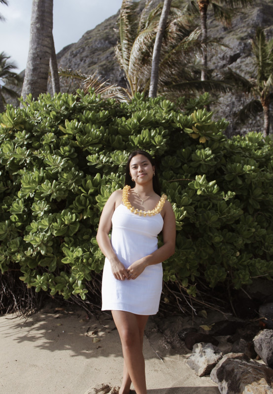 Girl standing on the beach with puakenikeni lei around her neck. Up against green plants and palm trees.