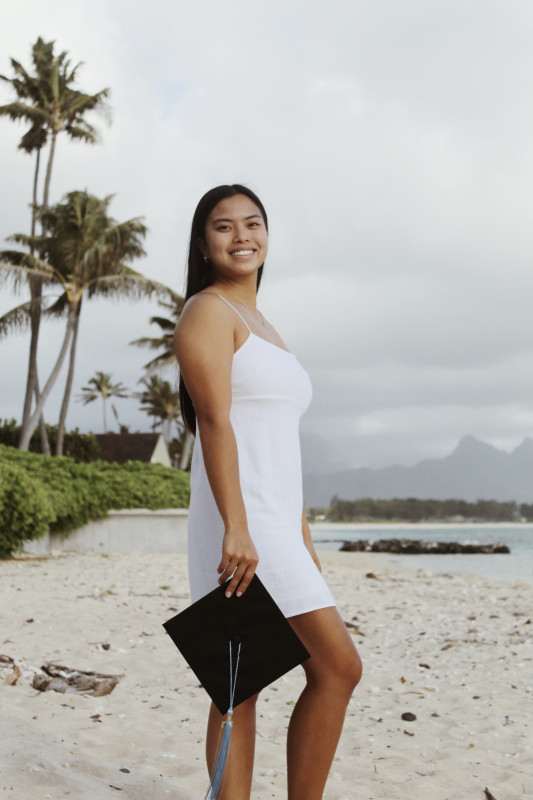 Girl standing on the beach with cap and tassel in hand. Smiling at the camera.