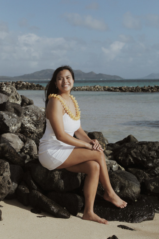 Girl sitting on a rock on the beach with a puakenikeni lei around her neck. Smiling at the camera.