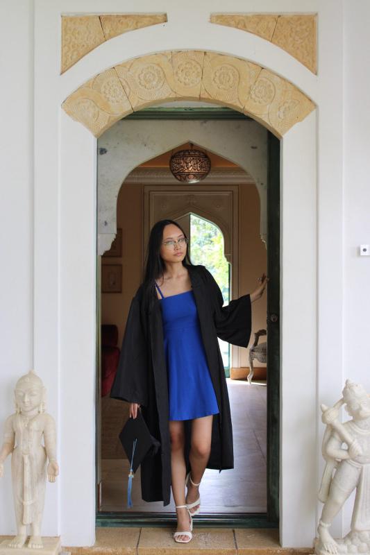 Graduate in a blue dress and graduation gown walking outside a door.