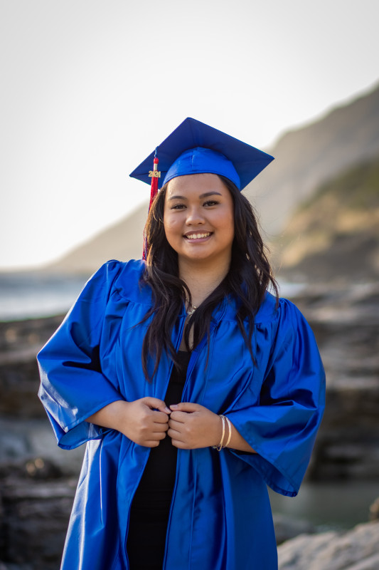 Blue graduation cap and gown, smiling with the sunset in the back.