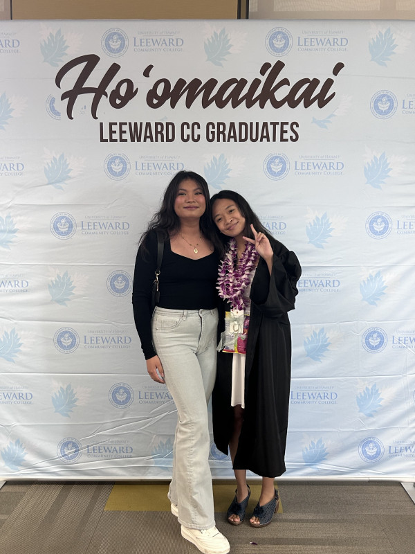 Two girls, one wearing a graduation gown, both posing in front of commencement backdrop