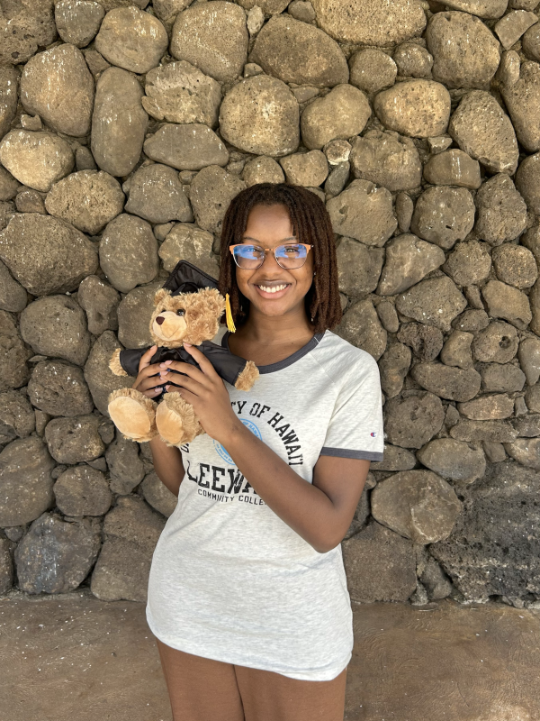 A black girl with short locs and glasses is wearing an LCC t-shirt and holding a graduation bear in front of a cobblestone wall on the LCC campus.