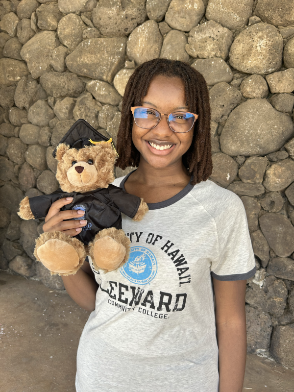 A black girl with locs wearing glasses and an LCC t-shirt and graduation bear is standing in front of a cobblestone wall while smiling to the viewer.