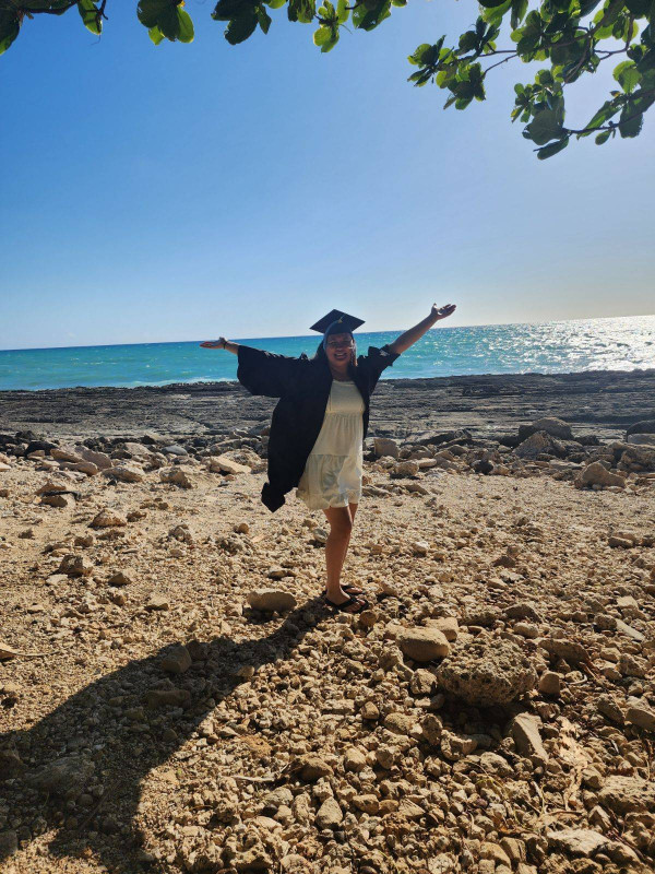 I'm posing in front of a rocky beach with the ocean in the backround and a tree hanging over. I'm wearing a white dress with my cap and gown.