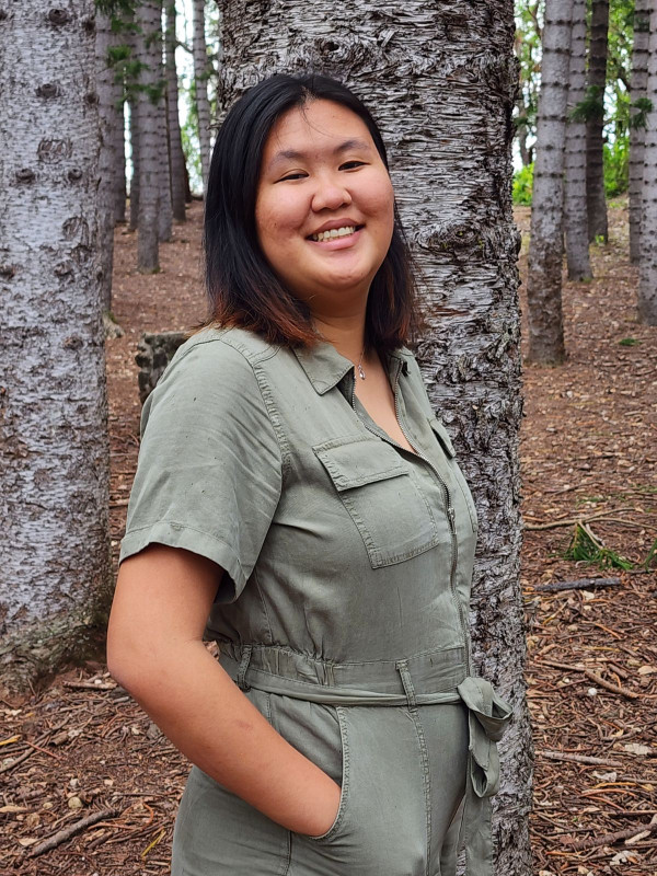 I'm leaning onto a tree in the middle of a forest, with my side profile facing the camera, I am wearing a green jumper.