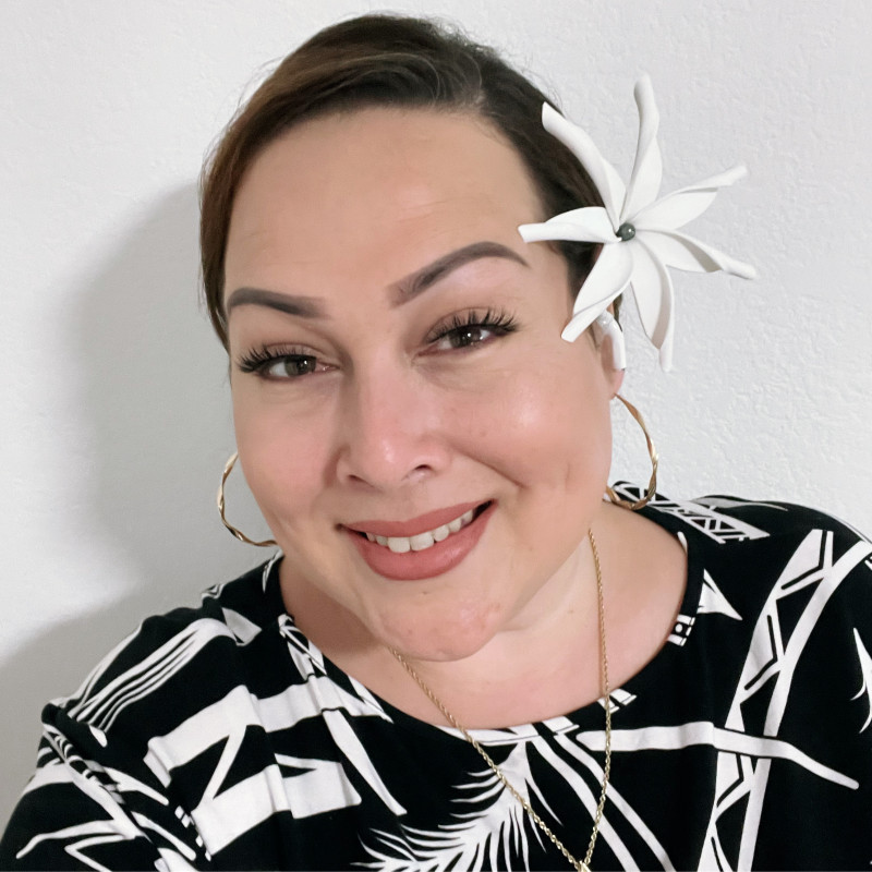 Fair complexion, slicked back hair bun, wearing a tiare flower on the left ear in Polynesian attire