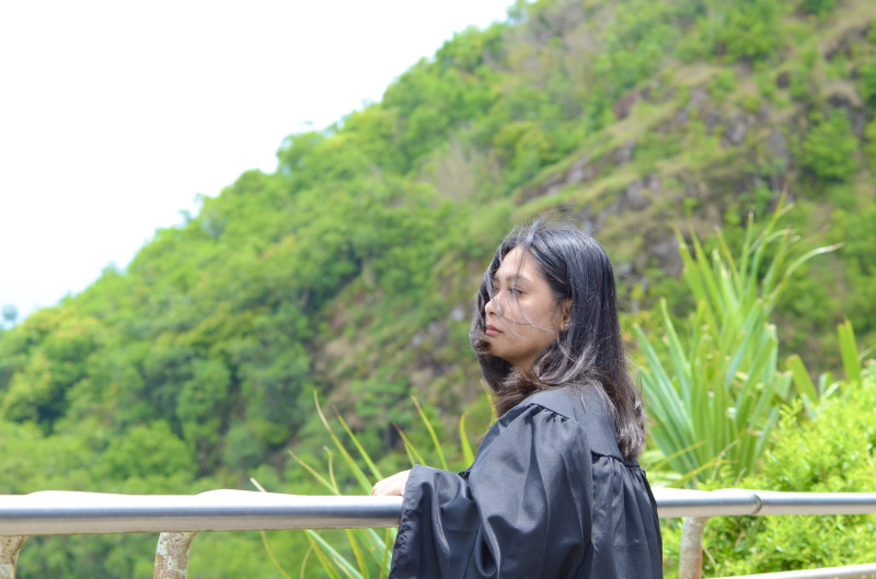 It’s a photo of me in my graduation gown, standing by a railing with lush green mountains behind me.
