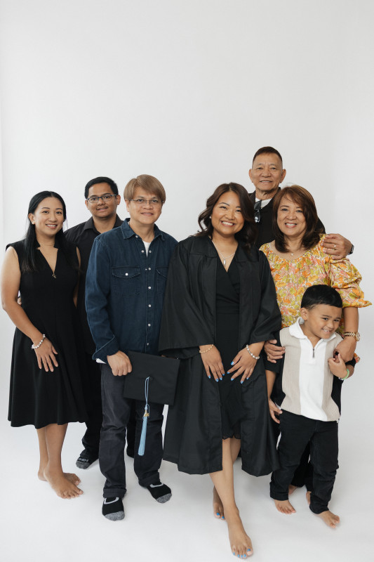 The image shows a joyful family graduation portrait. At the center stands a smiling woman in a black graduation gown, clearly the graduate, surrounded by six loved ones of various ages. Everyone looks happy and proud. The group includes a mix of men and women, a child in front who’s smiling playfully, and an older couple standing behind the graduate. They’re all barefoot or in socks, giving the photo a casual, intimate feel. The backdrop is clean and white, keeping the focus on the people and the emotion of the moment.