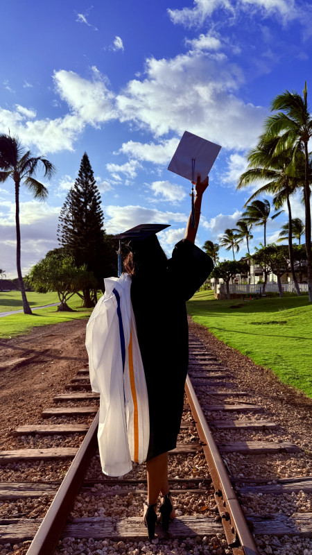 Caydence's back as she is wearing her black Leeward community college gown and cap while holding her Waipahu High School cap and gown.