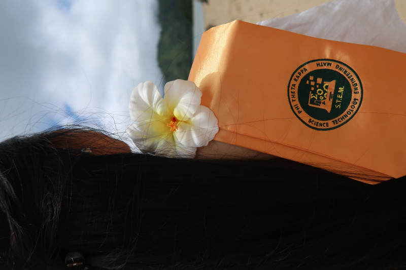 A picture of a girl from behind, with a white hibiscus resting on her shoulder and a gold stole indicating her membership in the Phi Theta Kappa National Honor Society and her STEM field of study.